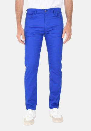 Uomo che indossa pantaloni slim fit blu brillante con bottone e cerniera frontale, abbinati a scarpe da ginnastica bianche e camicia bianca.