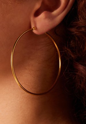Gouden creolen met een gladde, ronde vorm. Zichtbare sluiting nabij de oorlel, in contrast met de huidtint en krullend haar.