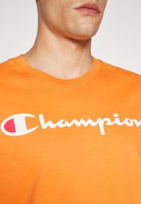 Orange Baumwoll-T-Shirt mit einem weißen "Champion"-Logo und einem roten Akzent. Rundhalsausschnitt mit kurzen Ärmeln. Glatte Textur.
