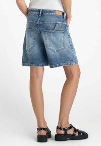 Shorts en denim bleu clair avec une coupe décontractée, présentant des poches arrière et un ourlet surpiqué. Associés à des sandales noires à lanières.