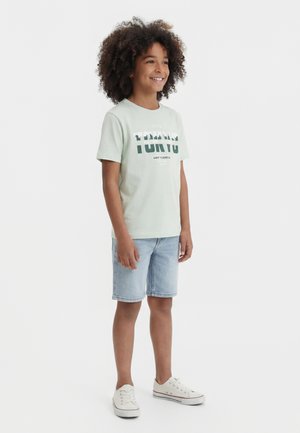 Enfant souriant aux cheveux bouclés portant un t-shirt graphique vert clair « Tokyo », un short en jean et des baskets blanches, debout sur un fond blanc.