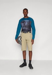 Herenfietskleding met een lange mouwen teal en donkergrijs shirt, khaki korte broek en zwarte sneakers. Houdt een grijze helm vast.