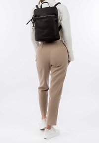Batoh vyrobený z texturované černé kůže, obdélníkového tvaru, se zipovou kapsou na přední straně a nastavitelnými popruhy. Nosený s béžovými kalhotami a bílými teniskami.