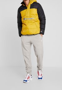 Vadderad jacka i gult och svart med hög krage, kombinerad med gråa sweatpants. Inkluderar en Champion-logotyp och sportiga skor i röd, blå och vit.