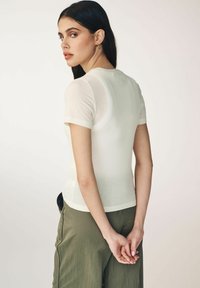 Leichtes, tailliertes Kurzarmoberteil in Creme mit glatter Textur und dezenten Rückennähten, kombiniert mit olivgrünen Hosen.