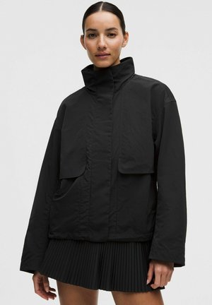 Sort, oversized jakke med høj krave, to forlommer og ribstrikkede manchetter. Glat tekstur, letvægtsstof, kombineret med et sort plisseret skørt.