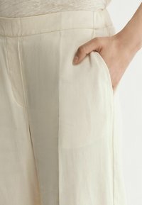 Ljust beige vida byxor med en slät textur, som har sidfickor och en ren midja. Subtil lyster synlig på tyget.