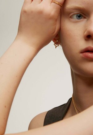 Pendientes de aro dorados con pequeñas puntas, adornados con una joya, llevados por una persona. Textura de piel suave y parte superior sin mangas de color marrón claro visible.