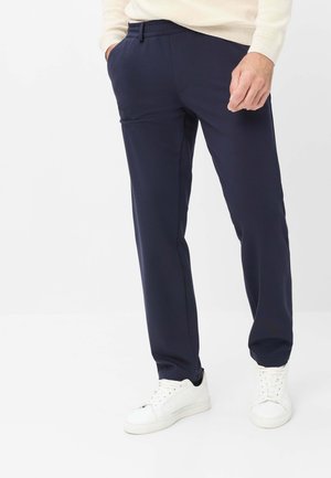 Mand iført marineblå bukser med lige ben, hvide sneakers og en cremefarvet langærmet top, står med den ene hånd i lommen.