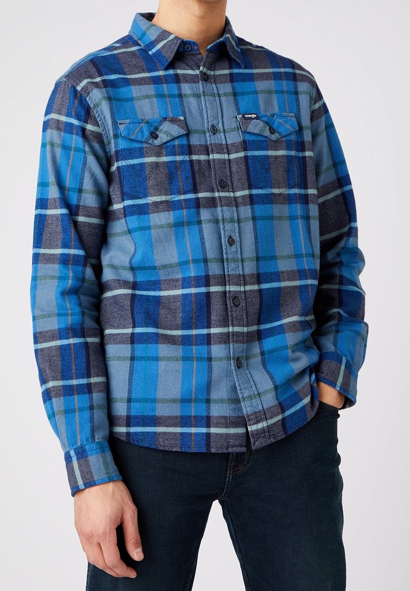 Chemise en flanelle à carreaux dans des nuances de bleu et de gris, dotée d'une patte de boutonnage, de deux poches poitrine et de manches longues avec poignets boutonnés.