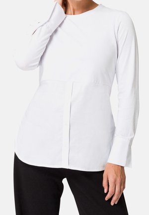 Camicia bianca a maniche lunghe con scollo rotondo, chiusura frontale con bottoni e cucitura asimmetrica sul petto; realizzata in tessuto liscio.