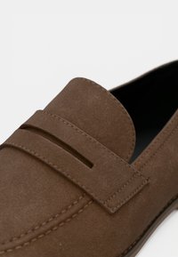 Καφέ σουέντ μοκασίνι με χαρακτηριστικό διπλό λουράκι, στρογγυλερή μύτη και αντίθετη ραφή κατά μήκος της σόλας και του άνω μέρους.