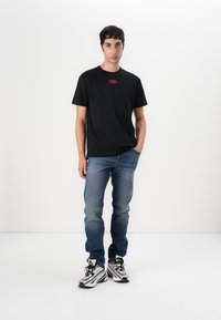 Zwarte katoenen T-shirt met een klein rood logo op de borst, gecombineerd met blauwe jeans en zwart-witte sneakers, staande tegen een witte achtergrond.