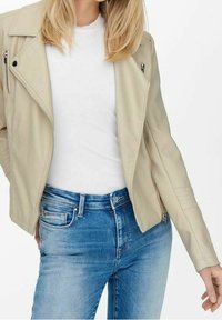 Beige läder-motojacka med silverblixtlås, figursydd design och texturerade axelackenter, kombinerad med en vit t-shirt och blå jeans.