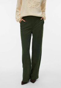 Person som har på seg mørkegrønne vide bukser, kremfarget strikkegenser med dekorative sømmer, hendene i lommene, og mørkebrune spisse sko.