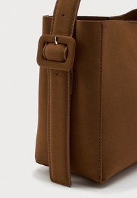 Braune Wildleder-Handtasche, die eine Nahaufnahme des verstellbaren Riemens mit quadratischer Schnalle und genähten Details zeigt.