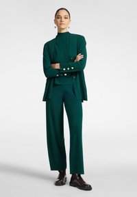 Conjunto de punto verde que presenta una blusa de cuello alto y un cárdigan largo con detalles dorados, combinado con pantalones de pierna ancha y botas negras hasta el tobillo.