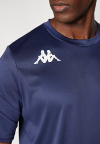 Camisola atlética azul-marinho com uma textura suave, apresentando um logótipo branco de duas figuras sentadas na parte superior esquerda da área do peito.