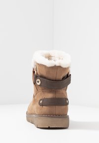 Bottines en suede marron avec une doublure en fausse fourrure blanche, une sangle marron foncé et un accent métallique, dotées d'une semelle en caoutchouc et d'un corps texturé.