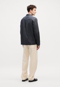 Camicia button-up grigia scura con una vestibilità rilassata, colletto e dettaglio a piega singola, abbinata a pantaloni color crema chiaro e scarpe nere.