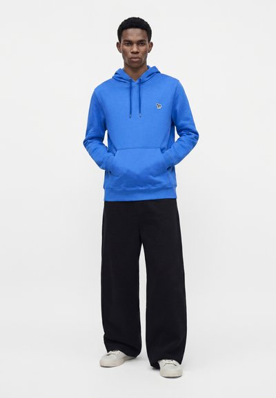 Homme debout portant un sweat à capuche bleu vif avec les mains dans la poche avant, pantalon noir à jambes larges et baskets blanches sur un fond uni.