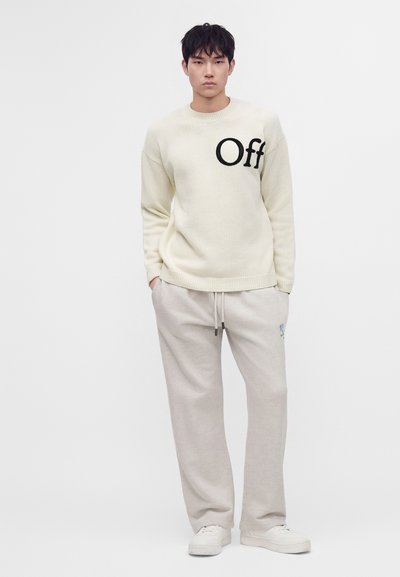 Jeune homme debout portant un pull crème avec le texte ""Off"" sur la poitrine, un pantalon de survêtement gris clair et des baskets blanches sur fond blanc.