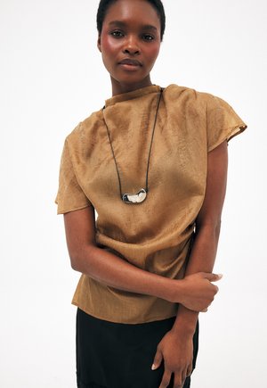 Kobieta z krótkimi włosami nosi brązową, teksturowaną bluzkę z krótkim rękawem, czarną spódnicę oraz długi czarny naszyjnik z srebrnym, abstrakcyjnym wisiorkiem.