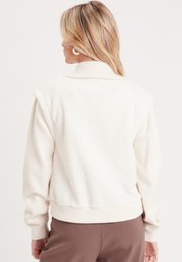 Chaleco de forro polar en color crema con un cuello acanalado, que presenta una textura suave y un diseño ajustado. Sin mangas, con un dobladillo inferior y puños de punto.
