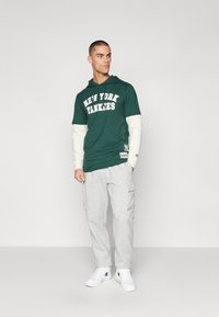 Zelená bavlnená mikina s kapucňou a potlačou "NEW YORK YANKEES", skombinovaná so šedými cargo teplákmi a bielymi teniskami. Neformálny športový strih.