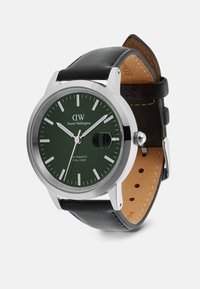 Daniel Wellington pulkstenis ar zaļu ciparnīcu, sudraba korpusu, trim rādītājiem, melnu ādas siksniņu un brūnu šuvju; automātiskais kustības mehānisms, 100m ūdensizturīgs.