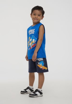 Canotta blu senza maniche con una grafica di Sonic the Hedgehog, abbinata a pantaloni corti blu scuro con un design simile. Sneakers in bianco e nero completano l'outfit.