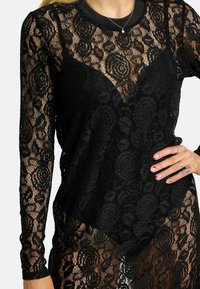 Fekete csipke felső bonyolult virágmintákkal, hosszú ujjakkal és mély V-kivágással. Áttetsző anyag, texturált felülettel.