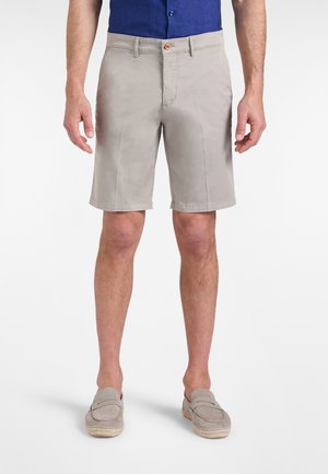 Uomo che indossa pantaloncini beige fino al ginocchio, camicia blu infilata e scarpe beige slip-on, in piedi su sfondo bianco.