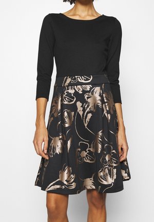 Femme portant une robe noire à manches trois-quarts et une jupe avec de grands motifs floraux métalliques.