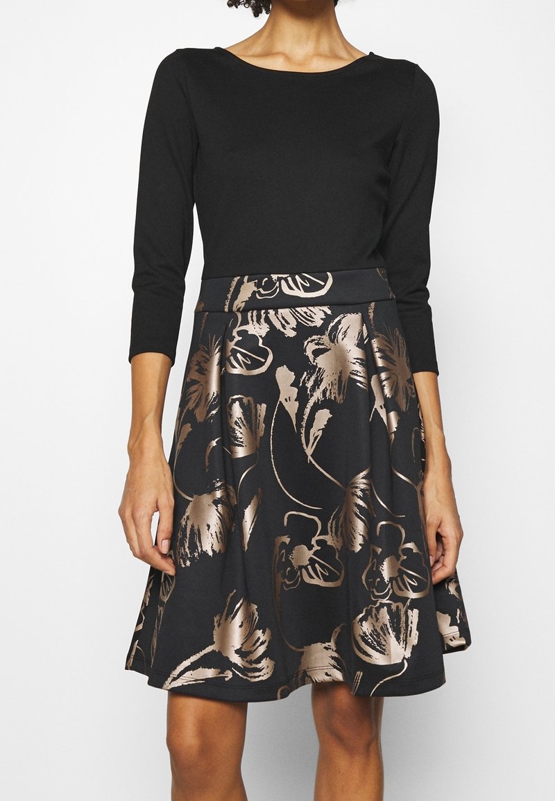 Femme portant une robe noire à manches trois-quarts et une jupe avec de grands motifs floraux métalliques.