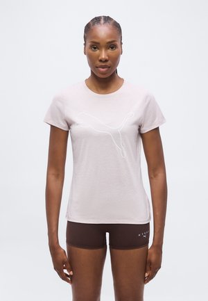 Femme portant un t-shirt Puma rose clair avec un logo blanc et un short Puma Hydro marron foncé, debout sur un fond clair uni.