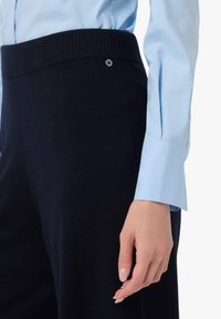 Pantaloni in maglia blu navy con cintura a coste, dotati di un dettaglio con bottone metallico. Abbina una camicia celeste con colletto.