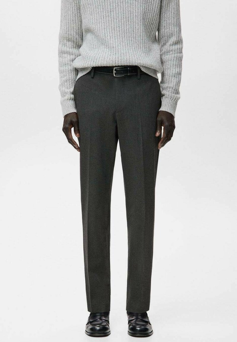 Homme portant un pantalon de costume gris foncé, une ceinture noire, des chaussures en cuir noires et un pull côtelé gris clair, debout devant un fond uni.