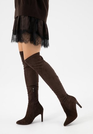 Vrouw die donkerbruine, suède laarzen met hoge hakken tot boven de knie draagt en een zwarte rok met kanten rand, staand tegen een witte achtergrond.