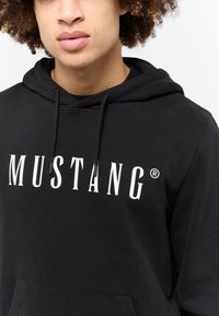 Felpa nera con logo bianco "MUSTANG", cappuccio con lacci e tasca a marsupio frontale. Realizzata in morbido tessuto testurizzato. Design casual.