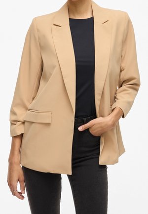 Woman wearing beige blazer over black shirt and dark jeans, with one hand in pocket and the other hanging down.