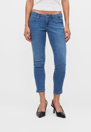 Femme portant un jean skinny bleu coupé au-dessus des chevilles et des talons slingback noirs, debout devant un fond blanc uni.