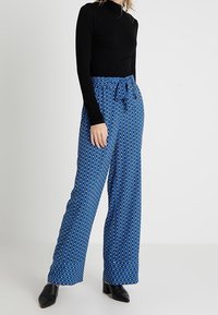 Blå mönstrade byxor med hög midja, knytbälte och vida ben. Matchas med en figurnära svart långärmad topp och svarta skor.