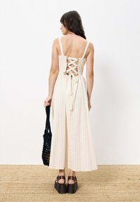 Cremesfarbene gestreifte Jumpsuit mit einem geschnürten Rückendesign und verstellbarem Band, kombiniert mit braunen Keilsandaletten und einer schwarzen geflochtenen Tasche.