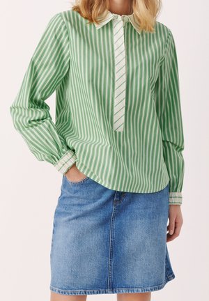 Femme portant un chemisier rayé vertical vert et blanc avec un col et des poignets blancs, associé à une jupe en jean bleue longueur genou.