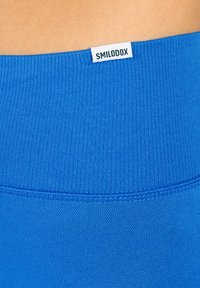 Ceinture de sport bleue avec texture côtelée, comportant une petite étiquette blanche portant la mention "SMILODOX" en haut. Matériau lisse sous la ceinture.