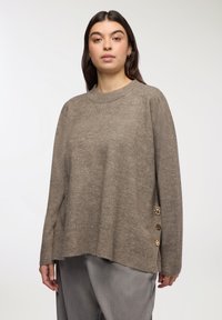 Maglione marrone oversize con orlo a coste, texture morbida e bottoni decorativi dorati di lato. Collo a giro e maniche lunghe.