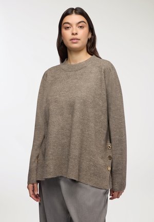 Maglione marrone oversize con orlo a coste, texture morbida e bottoni decorativi dorati di lato. Collo a giro e maniche lunghe.