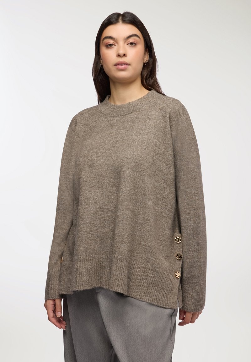 Maglione marrone oversize con orlo a coste, texture morbida e bottoni decorativi dorati di lato. Collo a giro e maniche lunghe.