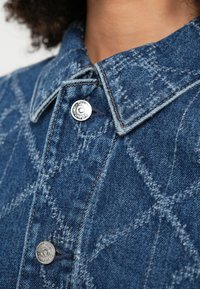 Niebieska denimowa koszula z kołnierzykiem, srebrnymi guzikami i pikowanym wzorem przeszyć. Tekstura tkaniny jest szorstka i widoczna.